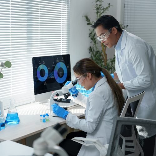 Scientists wearing lab coats and protective eyewear work in a laboratory. One uses a microscope while the other observes data on a computer screen, surrounded by scientific equipment