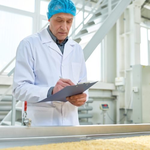Waist up portrait of senior factory worker  in food industry holding clipboard standing by conveyor belt, copy space